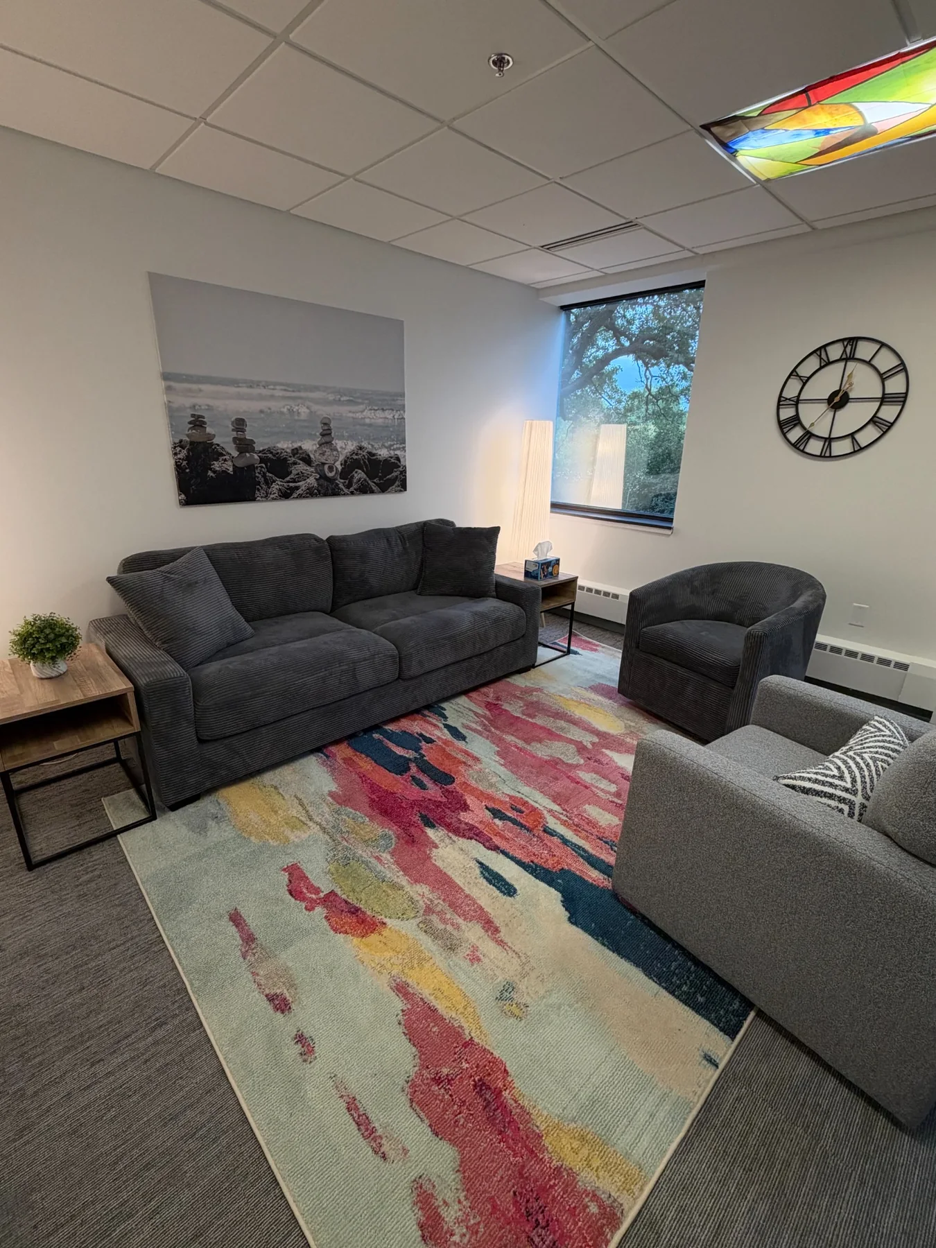 Therapy room with dark sofa, two chairs, colorful rug, stacked-stone wall art, and a wall clock