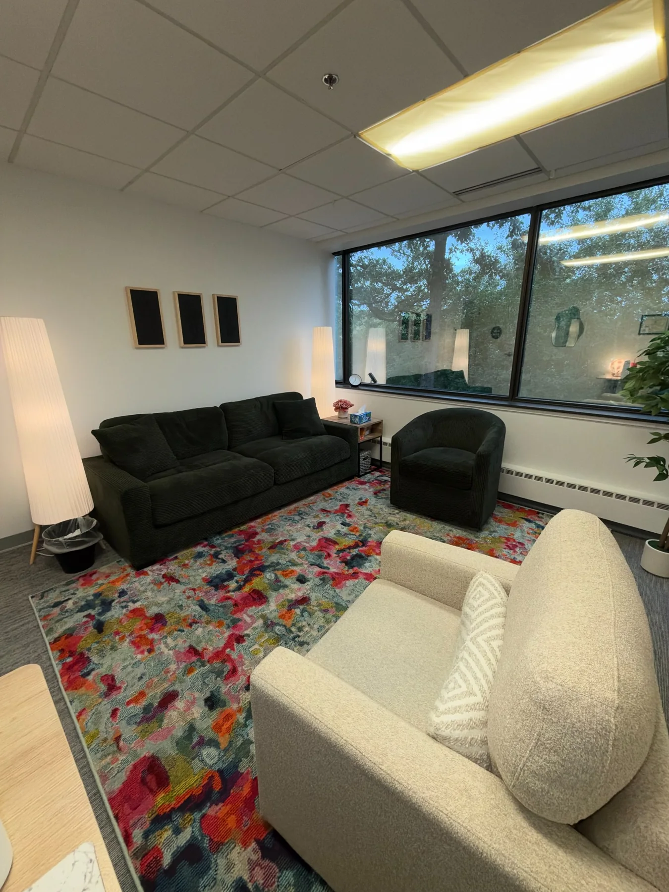 Therapy room with dark sofa, cream chair, colorful rug, large windows, and soft floor lamps